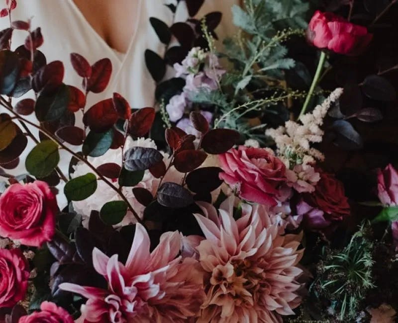 A close-up view of a person in a white dress holding a large and colorful bouquet of flowers. The bouquet features a mix of pink roses, dahlias, dark foliage, and other greenery, creating a rich and vibrant display.