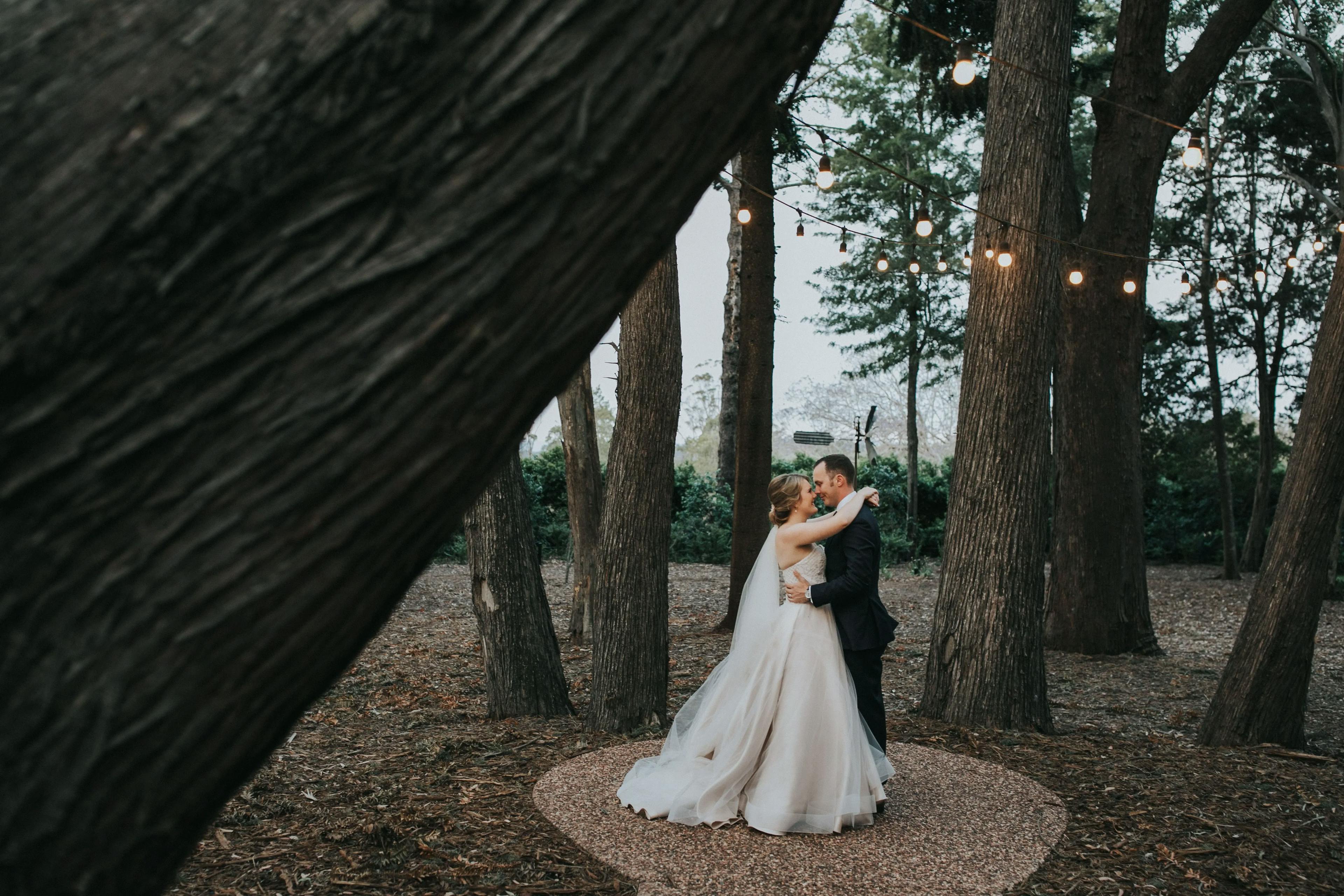 A bride and groom share a romantic embrace outdoors in a forested area. The couple stands on a circular path, framed by tree trunks, beneath string lights that hang above them, creating a magical atmosphere. The bride wears a flowing wedding dress, and the groom is in a suit.
