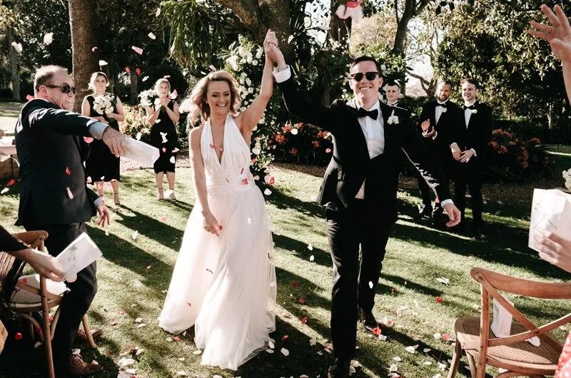 A bride in a white dress and a groom in a black tuxedo joyfully walk down an outdoor aisle, with their hands raised and smiling faces. Guests cheer and throw flower petals as the couple passes by. The scene is set in a lush garden with trees and greenery.