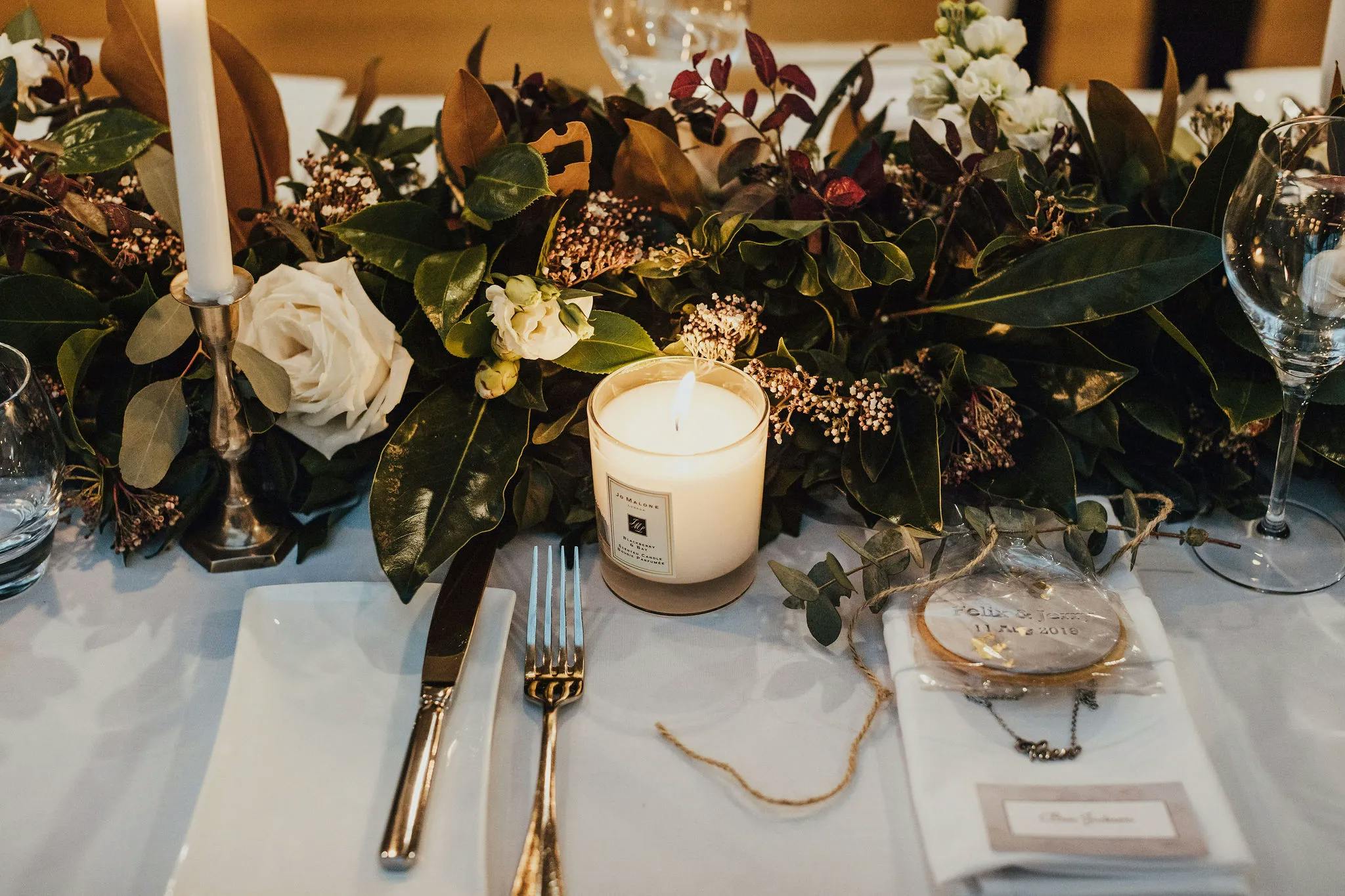 A beautifully decorated table set for a formal meal. There are elegant place settings with a knife, fork, and napkin, a lit candle, and lush green and white floral arrangements. Wine glasses and a name card are also present, adding a touch of sophistication.