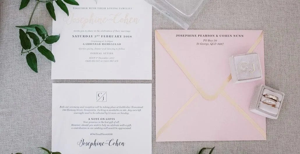 A wedding invitation set includes an RSVP card, an invitation card, and a card about gifts, all laid out on a light gray surface with green leaves. To the right, there's a pink envelope, a return address card, and a clear box containing two rings.
