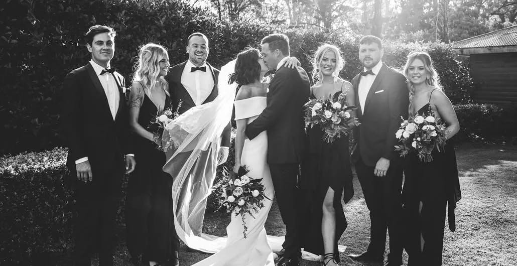 A black and white photo of a wedding party. The bride and groom are kissing in the center, surrounded by groomsmen in suits and bridesmaids in dresses. The bride holds a bouquet and wears a long veil, and everyone is smiling, standing outdoors.