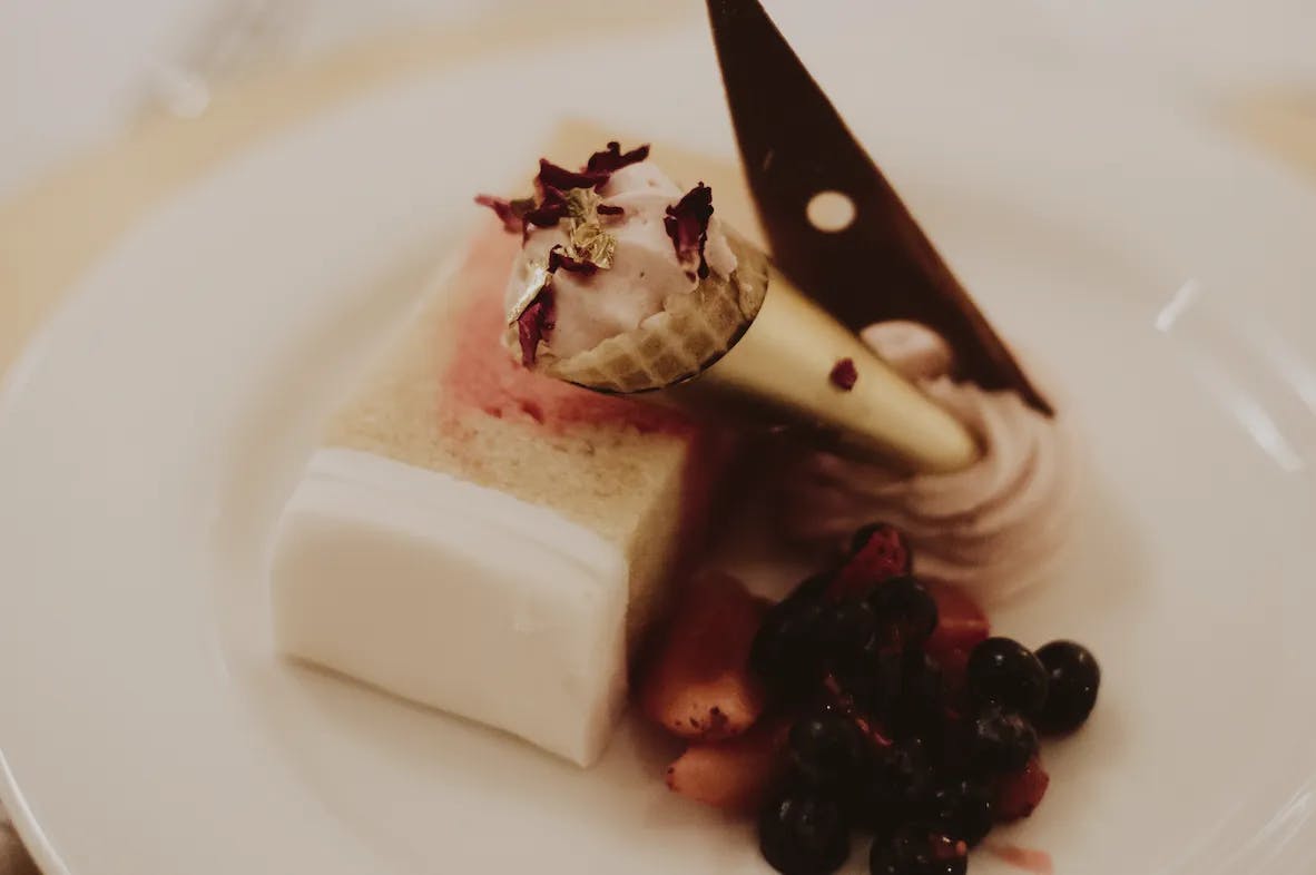 A gourmet dessert, featuring a slice of layered cake with white icing, topped with a delicate mini cone filled with cream and garnished with rose petals. Beside it is a small serving of mixed berries and a dark chocolate decoration, all presented on a white plate.
