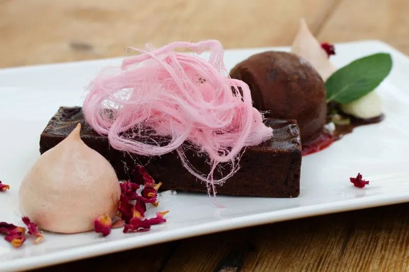 A neatly arranged dessert plate featuring a chocolate brownie topped with a nest of pink cotton candy. Accompanying the brownie is a scoop of chocolate ice cream, a light pink meringue, edible flowers, and green leaves on a white rectangular plate.