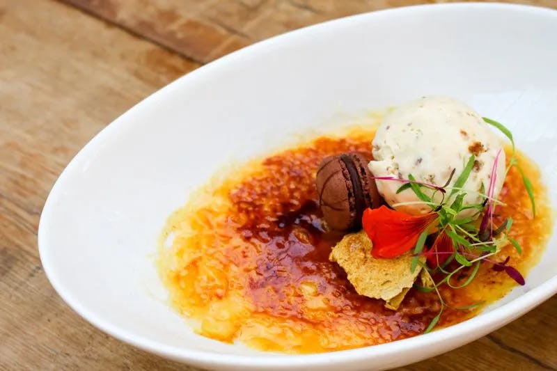 A white oval dish contains a crème brûlée topped with a scoop of vanilla ice cream, a chocolate macaron, and a decorative edible flower with microgreens. The burnt sugar topping on the crème brûlée is caramelized to a golden brown.
