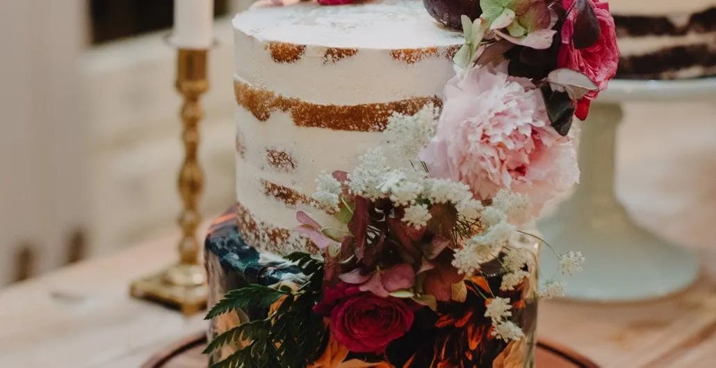 A semi-naked cake topped with pink, white, and deep red flowers. There are green foliage and berries garnishing the cake, which rests on a glass stand. A white candle in a brass holder is visible in the background. The setting suggests a rustic or elegant decor.