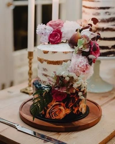A rustic two-tiered semi-naked cake adorned with an assortment of vibrant flowers and greenery. The cake is displayed on a wooden board, with candles and another cake visible in the background. A cake knife and server are placed beside it.