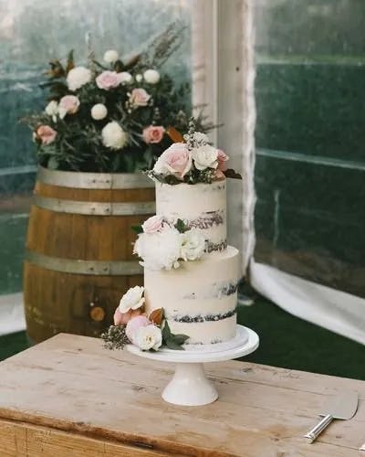 A two-tier white frosted cake adorned with pink and white flowers on a white pedestal, displayed on a wooden table. In the background, there is a wooden barrel decorated with similar flowers, set in a tented outdoor venue.