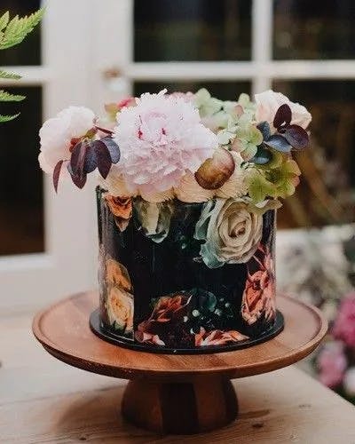 A decorative cake adorned with floral designs on its sides rests on a wooden pedestal. The cake is topped with realistic-looking flowers, including pink peonies and roses, along with green leaves, creating an elegant and sophisticated appearance.