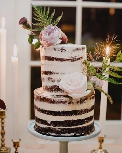 A two-tier naked cake with layers of chocolate and white frosting is decorated with pink roses, greenery, and a small twig. The cake is placed on a light blue cake stand, surrounded by white candles in brass holders, against a window-paneled background.