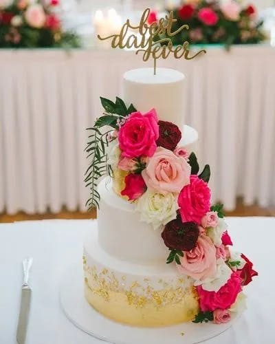 A three-tier white wedding cake adorned with a cascade of red, pink, and white roses. The top tier has a gold topper that reads "Best day ever." The lower tier is decorated with gold leaf. The background features a table with a floral arrangement.