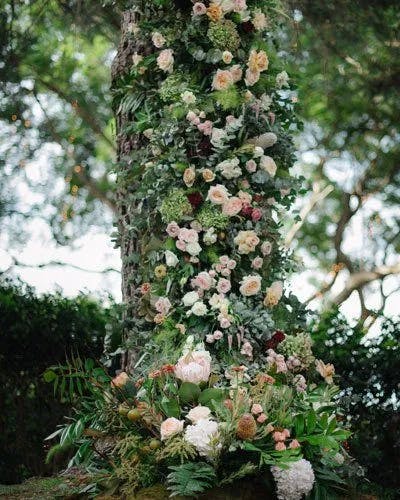 A large tree trunk is adorned with a beautiful arrangement of flowers and greenery. The display features various types of flowers in shades of pink, white, and green, with lush foliage cascading down the trunk and blooming at the base. The background is filled with trees and greenery.