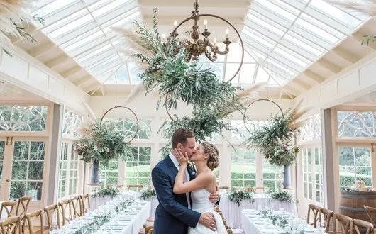 A couple dressed in wedding attire shares a kiss in a beautifully decorated, sunlit conservatory. The room features elegant chandeliers adorned with lush green foliage and long banquet tables set for a reception, positioned beneath a glass roof.