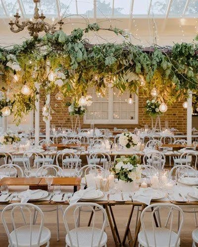A festive banquet hall decorated with lush greenery and hanging string lights adorned with Edison bulbs. The tables are set with white plates, glasses, and floral centerpieces. White chairs surround the tables, and a chandelier hangs above the setup.