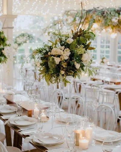 Elegant event setup featuring long tables adorned with white tablecloths, green and white floral centerpieces, and softly lit candles. The background showcases twinkling string lights and additional hanging greenery, creating a romantic ambiance.