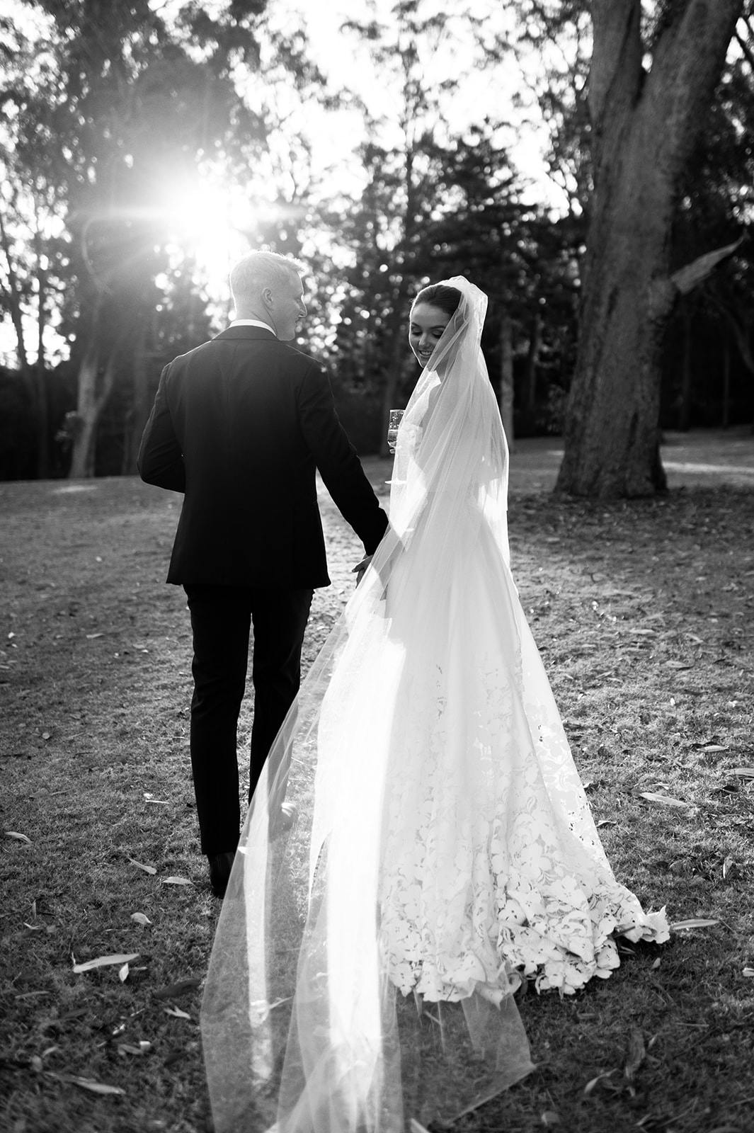 A black and white photograph of a newlywed couple walking hand-in-hand in a park. The bride wears a long, flowing dress with a veil, and the groom is dressed in a suit. The sunlight filters through the trees, casting a soft glow on the couple.