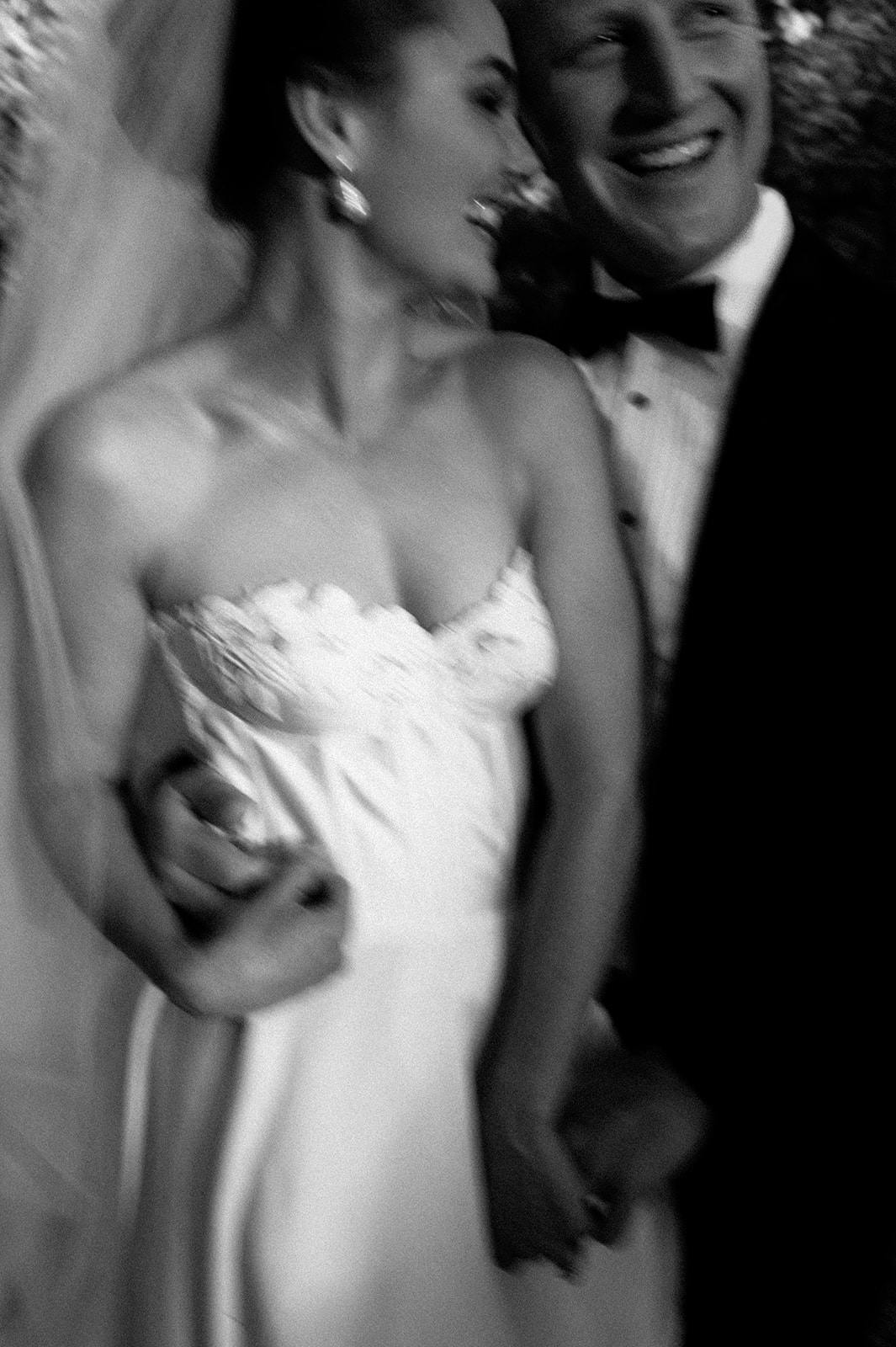A black and white photo of a bride and groom. They are standing close together, smiling and laughing. The bride is wearing a strapless wedding gown and veil, while the groom is in a tuxedo with a bow tie. The image is slightly blurred, capturing a candid moment.