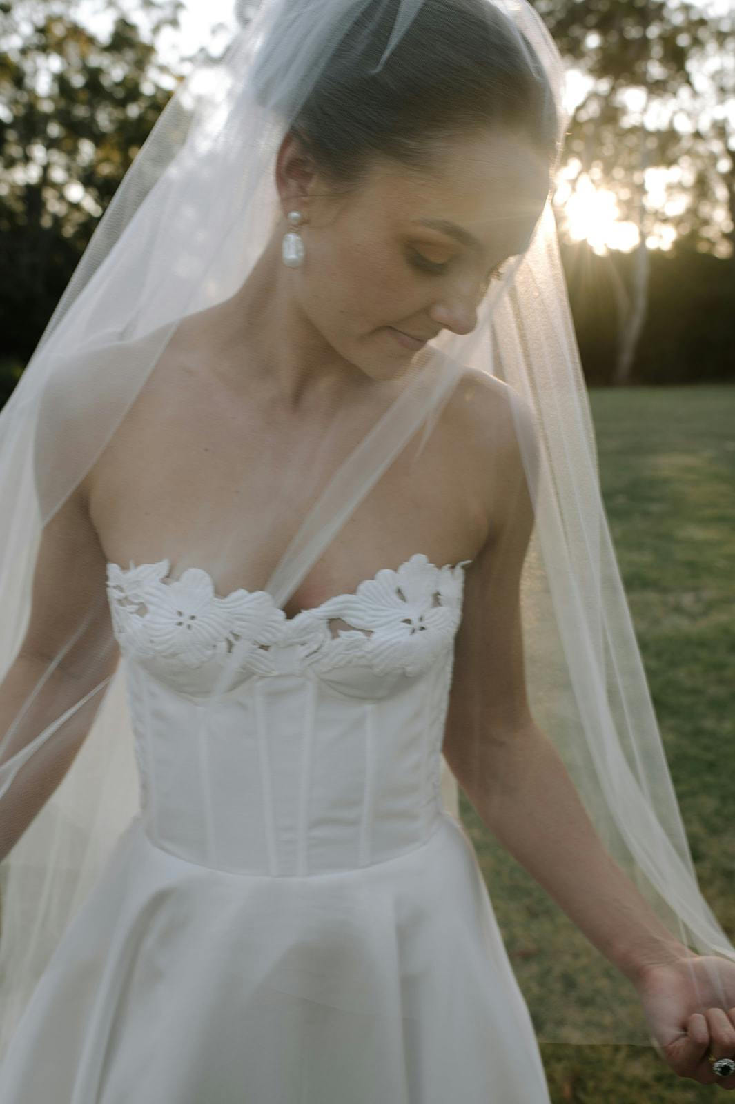 A bride wearing a strapless white wedding gown with floral lace details and a long veil stands outdoors. She looks down thoughtfully, with sunlight filtering through trees in the background.