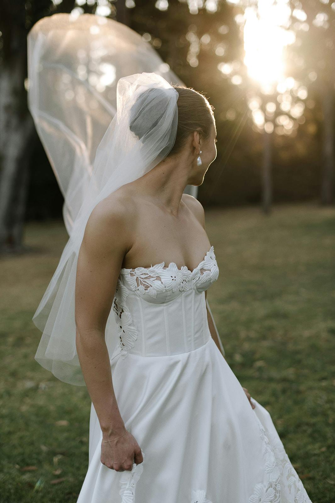 A bride in a strapless white wedding dress and long veil stands outdoors, looking to her left. The sunlight filters through the trees in the background, casting a soft glow. She is holding the skirt of her dress, and the scene is serene and picturesque.