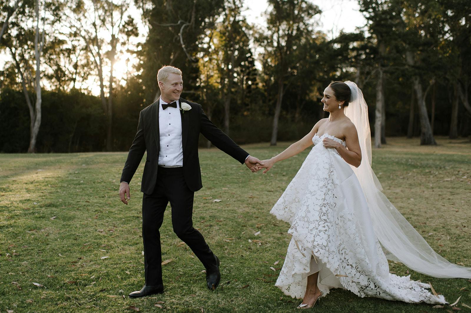 A couple walks hand-in-hand on a grassy field at sunset. The groom wears a black tuxedo with a white rose boutonniere, while the bride wears a strapless white lace wedding dress and a long veil. Both are smiling as they look at each other. Trees are in the background.