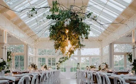 A bright and elegant wedding reception venue with glass ceilings and walls, adorned with strings of fairy lights and hanging greenery. The space is filled with white chairs and tables decorated with floral centerpieces, creating a romantic and sophisticated atmosphere.