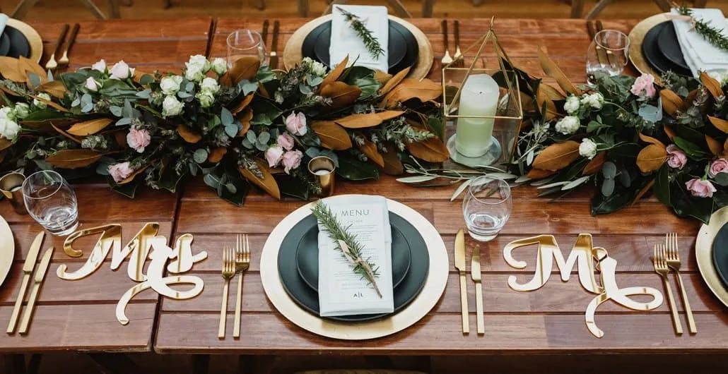 A beautifully set wooden table with elegant gold and black tableware for a wedding. Lush floral green and white centerpieces, including leaves and candles, run along the table. Wooden "Mr." and "Mrs." signs mark the couple's places at the head of the table.