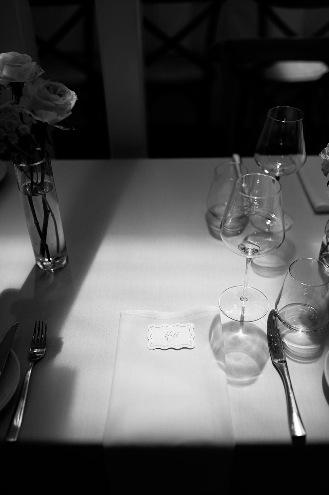 Black and white photo of a neatly set table with two vases of flowers, multiple empty wine glasses, and a place card reading "Mr" on a napkin. The scene is illuminated by soft natural light, casting shadows on the table.