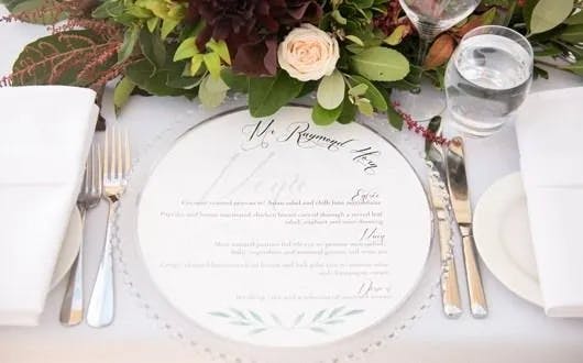 A beautifully arranged table setting with a white plate featuring elegant calligraphy text placed under a glass charger. The setting includes a folded white napkin, silver cutlery, a glass of water, and a lush, colorful floral centerpiece.