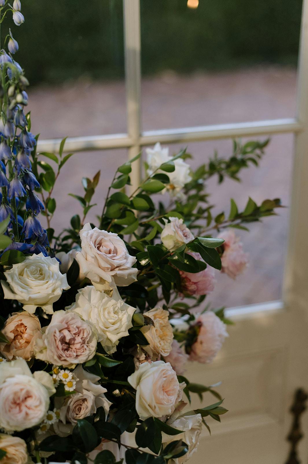 A floral arrangement with pastel roses, peonies, and greenery is positioned in front of a glass door. The soft pink, white, and peach flowers are complemented by green leaves and a few tall, blue flowers, with a brick patio visible outside through the glass.