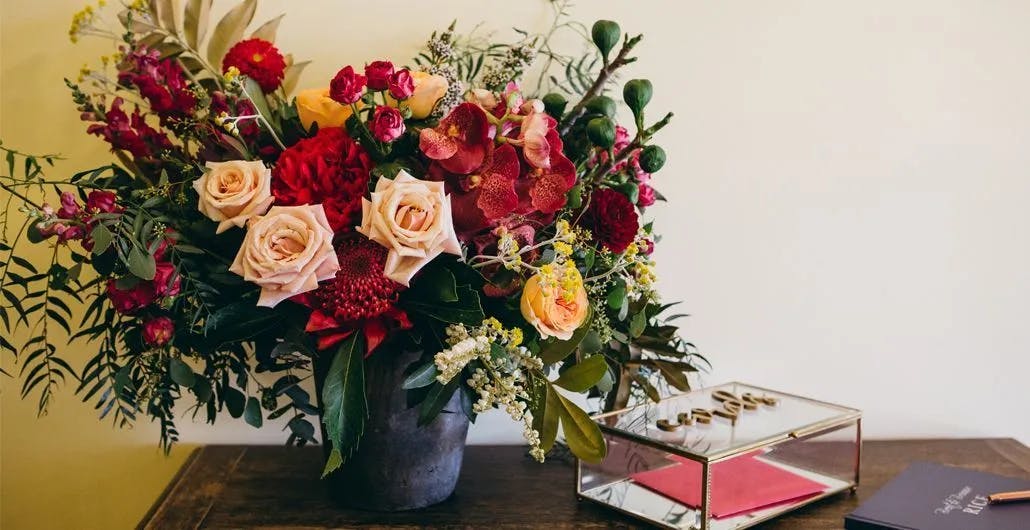 A vibrant floral arrangement featuring red and peach roses, assorted greenery, and other colorful flowers in a black pot sits on a wooden table. To the right, there is a glass box with gold rings inside and a closed book on the table.