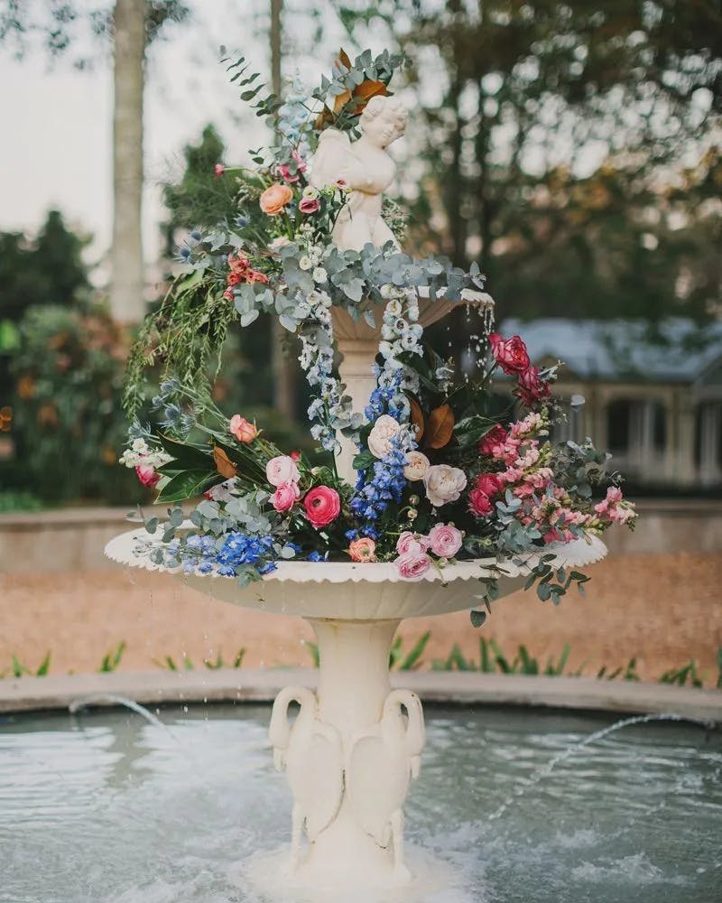 A beautiful white stone fountain with water flowing over the tiers is adorned with vibrant, fresh flowers in pink, blue, and white. Greenery and a small statue of an angel embellish the top, set against a lush garden backdrop.