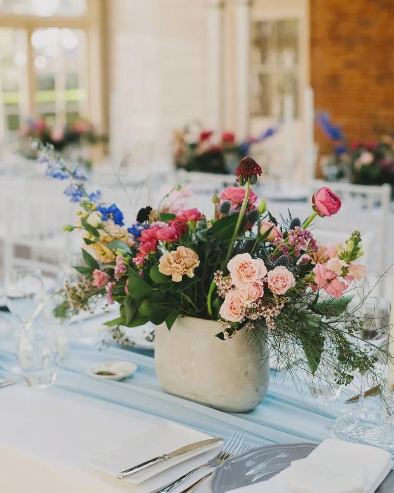 Elegant table setting with a centerpiece featuring a mix of vibrant and pastel flowers in a ceramic vase. The table is adorned with white linens, silver cutlery, wine glasses, and a subtle light blue table runner, situated in a bright, airy venue.