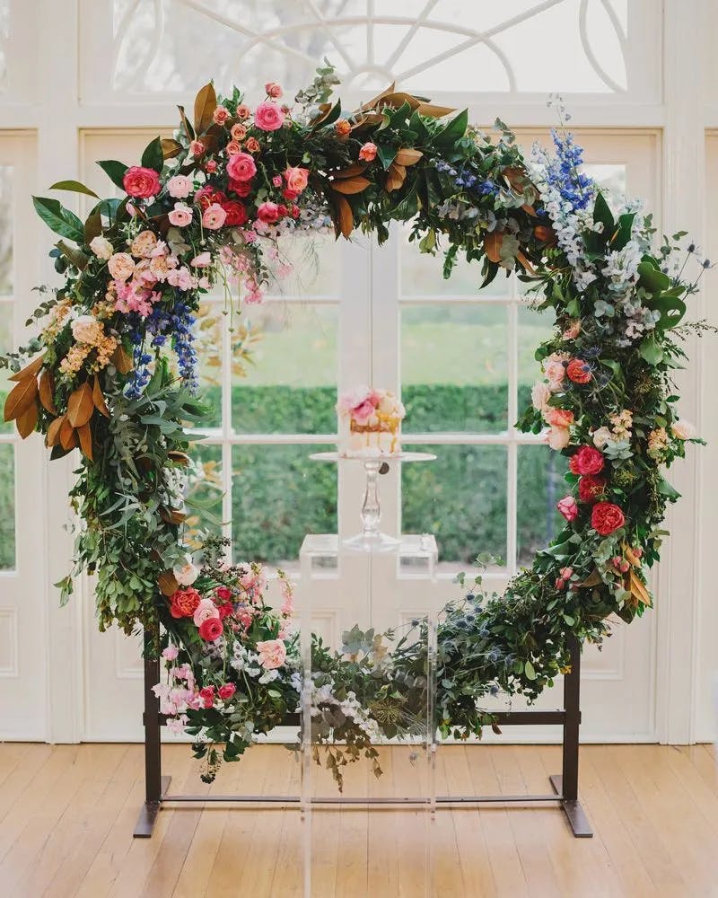 A large circular floral arrangement is adorned with vibrant flowers in shades of pink, red, white, blue, and green foliage. It stands in front of French doors with glass panels, allowing a view of a lush garden outside. At the center, a small vase with pink flowers is displayed.