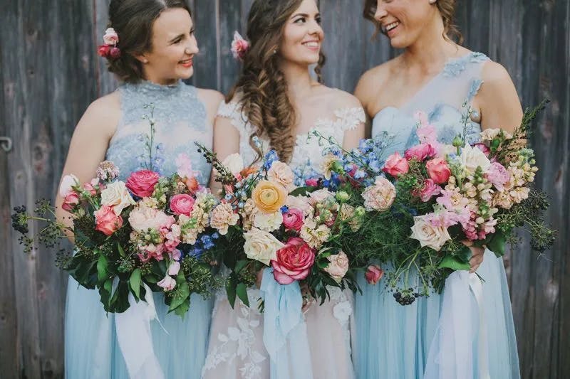Three women wearing light blue dresses, with the central figure in a white gown, stand smiling and holding large, colorful bouquets of roses and other flowers. They are in front of a wooden wall, creating a rustic backdrop. Two of the women have flowers in their hair.
