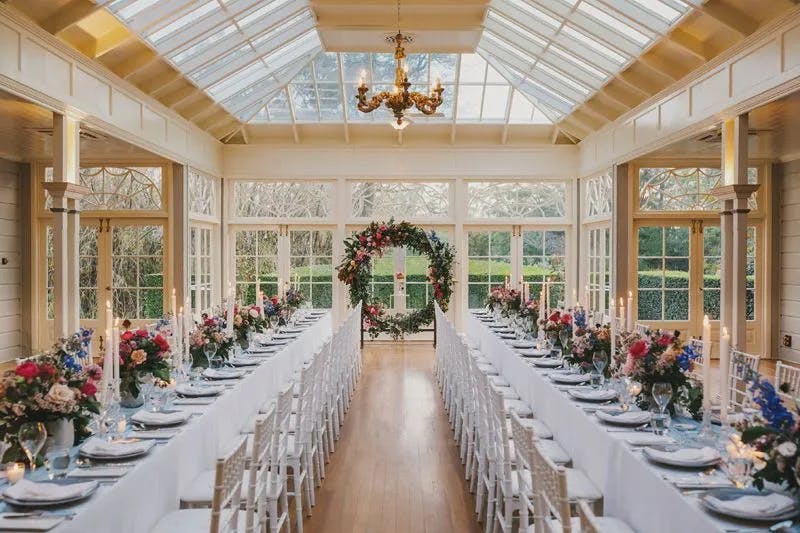A well-lit, elegant wedding venue with two long dining tables adorned with colorful floral centerpieces, glassware, and place settings. The tables are aligned parallel under a glass ceiling, leading to a large floral arch at the end of the room.