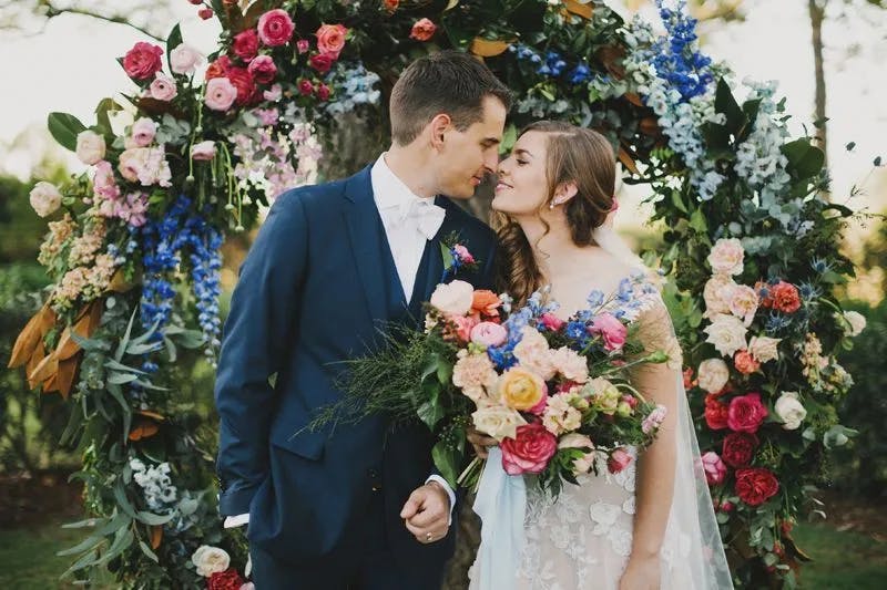 A bride and groom stand in front of a large arch adorned with colorful flowers. The groom, in a navy suit and white bow tie, gazes at the bride, who wears a lace wedding dress and holds a vibrant bouquet. They stand close, nearly touching foreheads.