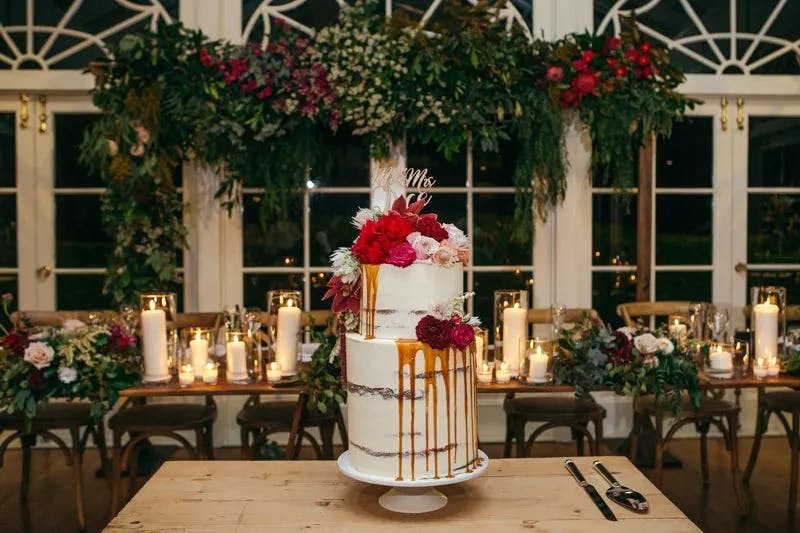 A white, semi-naked wedding cake adorned with red, pink, and white flowers and a "Mr & Mrs" topper stands on a wooden table. The cake features a caramel drip design. In the background, lit candles and a lush greenery garland decorate the windowed wall.