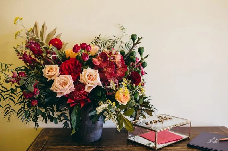A vibrant bouquet of flowers featuring pink roses, red dahlias, and mixed greenery in a dark vase is placed on a wooden table. Beside the vase is a glass box with gold edges containing a pink and red notebook or card, and a closed dark blue book.
