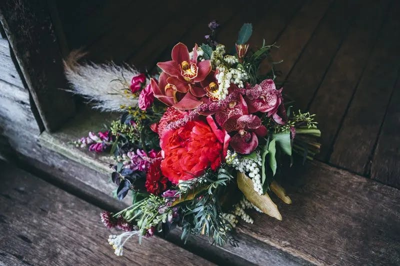 A vibrant bouquet featuring red peonies, pink orchids, and various other colorful flowers and greenery, set against a weathered wooden surface. The arrangement includes different textures and shades, creating a rich and rustic aesthetic.