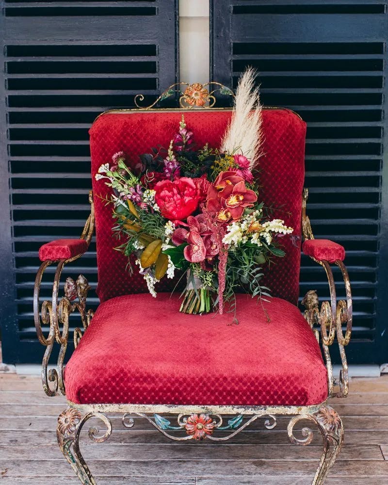 A rustic ornate chair with a red cushioned seat and backrest is centered in front of dark shutters. On the seat, a vibrant bouquet of flowers, including red and pink blooms, green foliage, and white accents, is on display. The chair sits on a wooden deck.