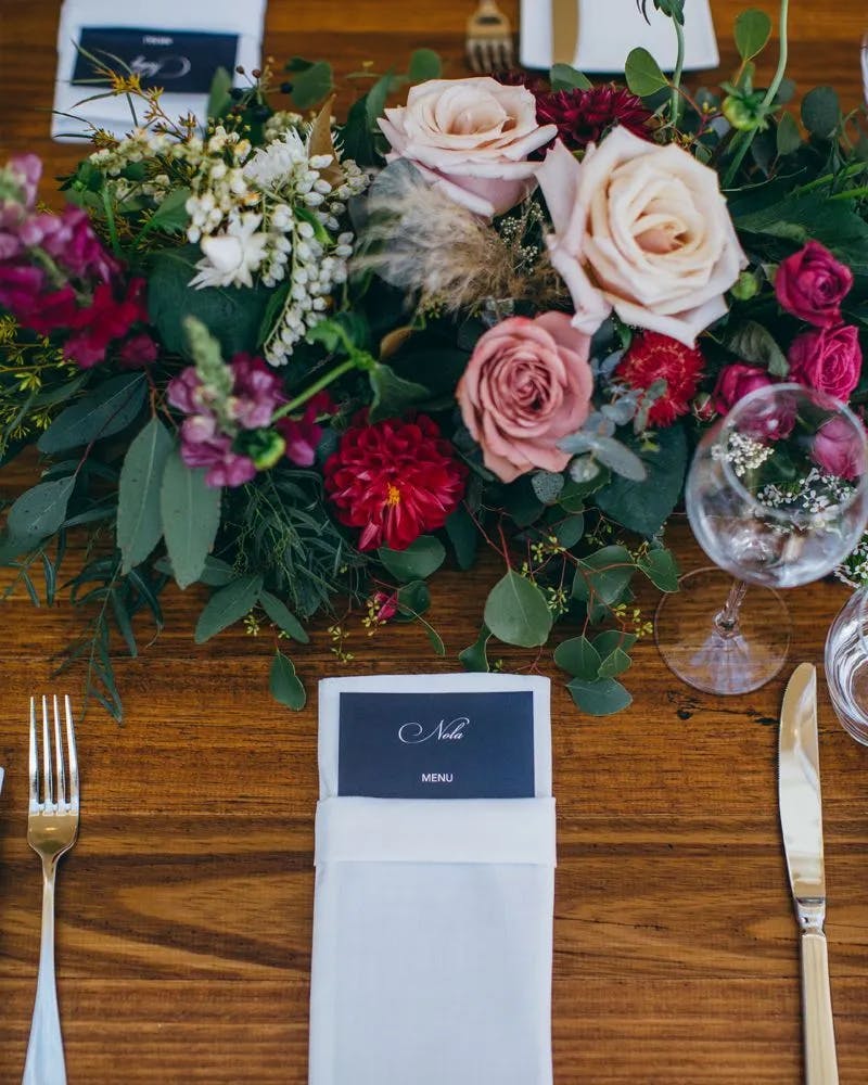 A beautifully set wooden table adorned with a lush floral arrangement featuring pink roses and various greenery. A neatly folded white napkin holds a navy menu card. Nearby are a wine glass, fork, and butter knife, creating an elegant dining setup.