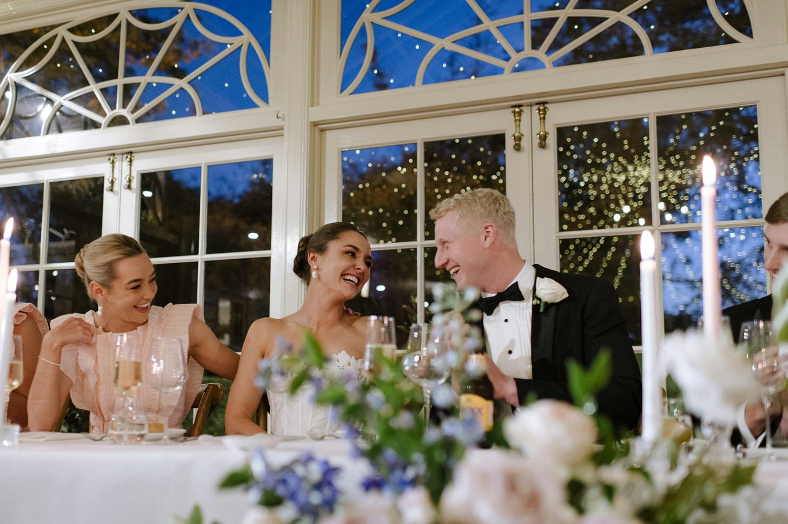 A bride and groom, dressed in formal wedding attire, sit at a beautifully decorated table laughing and engaging with bridesmaids and guests. The background features large windows with an evening view and twinkling lights. The scene is joyful and elegant.