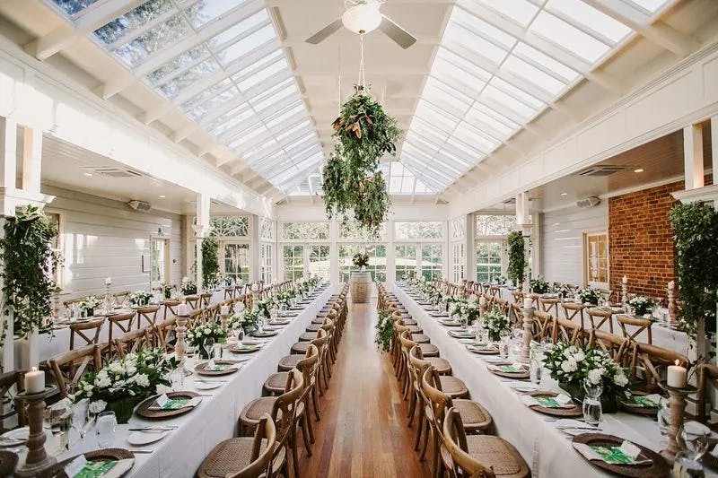 A beautifully decorated banquet hall with a glass ceiling, featuring long, parallel wooden tables adorned with white tablecloths, green and white floral arrangements, and candle holders. The hall is filled with natural light, creating an elegant and airy ambiance.