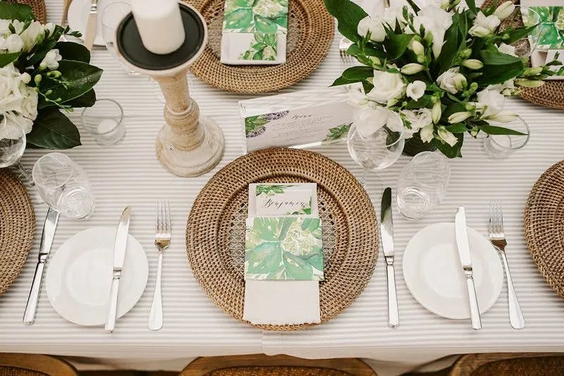A beautifully arranged table setting with wicker placemats, white plates, silver cutlery, and green-themed menus. White flowers and greenery are used as centerpieces, and a white candle on a rustic holder adds elegance to the table.