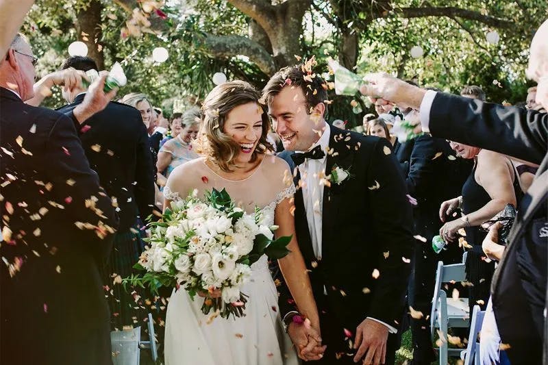 A joyful bride and groom walk down the aisle outdoors, smiling and holding hands as guests throw flower petals over them. The bride holds a large bouquet of white flowers, while the groom wears a black suit and bow tie. Trees and decorations can be seen in the background.