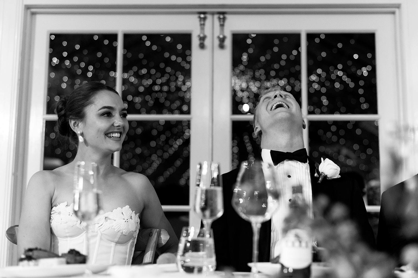 A black and white photo captures a joyous moment between a bride and groom. The bride, in a strapless dress, smiles warmly, while the groom, in a tuxedo, leans back laughing. The background features twinkling lights, creating a magical atmosphere.