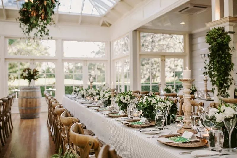 A beautifully set long dining table is adorned with white linens, greenery, white flowers, and candles. Wooden chairs surround the table in a bright room with large windows and ample natural light. Lush plants hang from the ceiling, enhancing the elegant atmosphere.