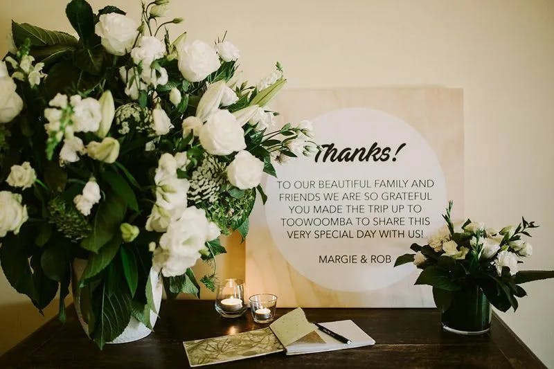 A decorated table with two floral arrangements of white flowers and greenery. There are candles, a small notebook, and a large card that reads, "Thanks! To our beautiful family and friends, we are so grateful you made the trip up to Toowoomba to share this very special day with us! Margie & Rob.