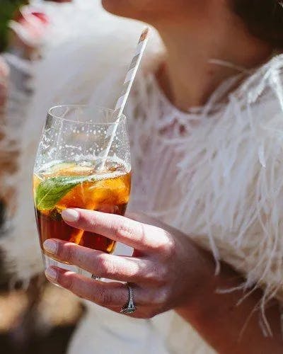A person holding a glass filled with iced tea garnished with lime and a striped straw. The person is wearing a white, feathered outfit, and a diamond ring is visible on their finger. The background is softly blurred.
