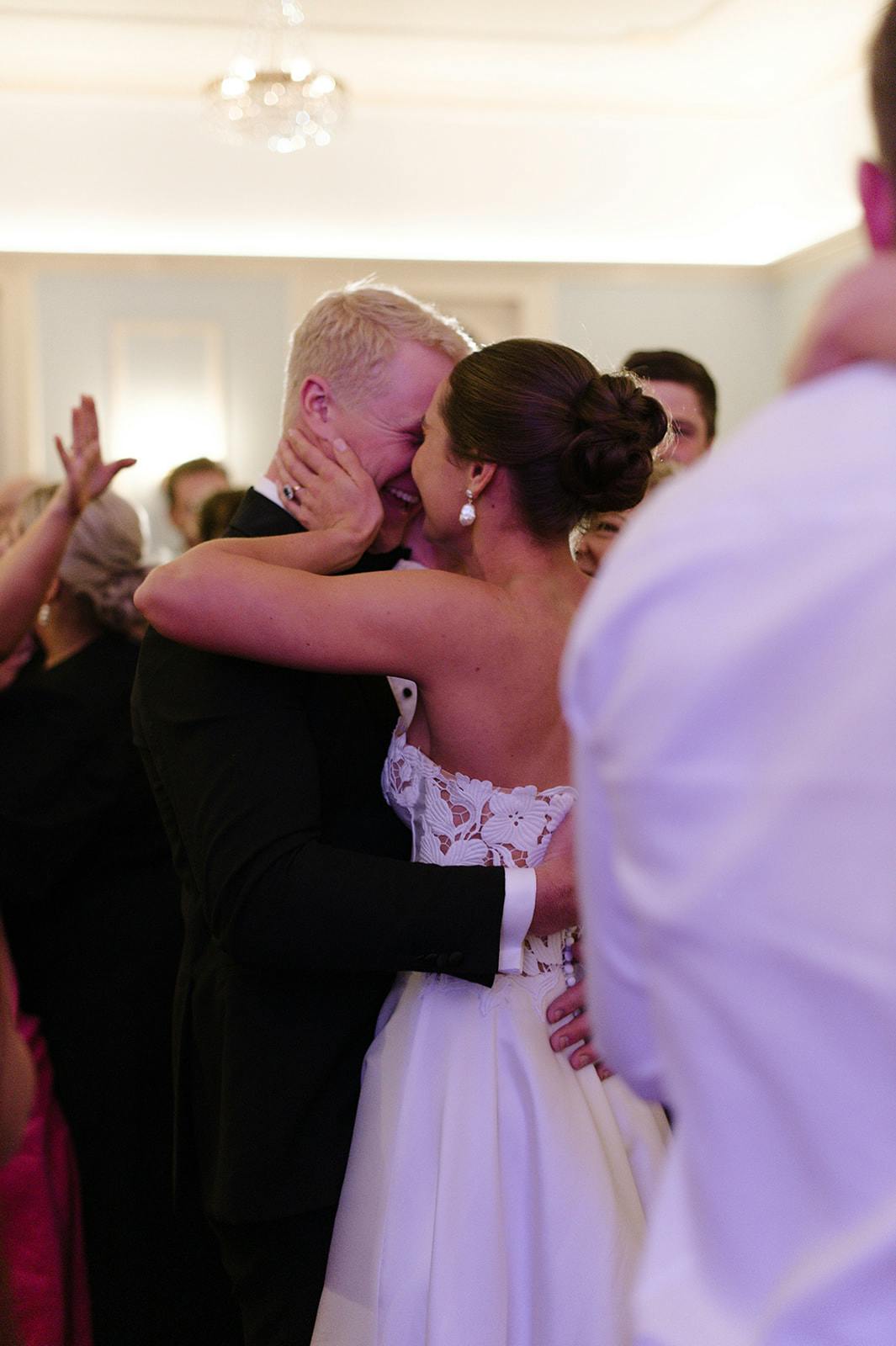 A newlywed couple shares a joyful embrace during their wedding reception. The bride, dressed in a white gown with lace details, has her arms wrapped around the groom, who is in a dark suit. They are surrounded by cheering guests, capturing a moment of celebration and love.